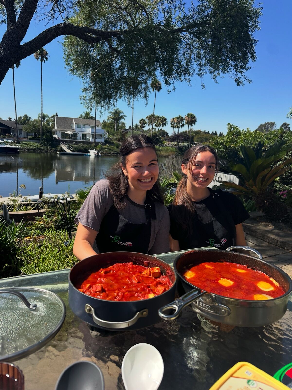 Two guests behind pans of shakshuka at a table by the water