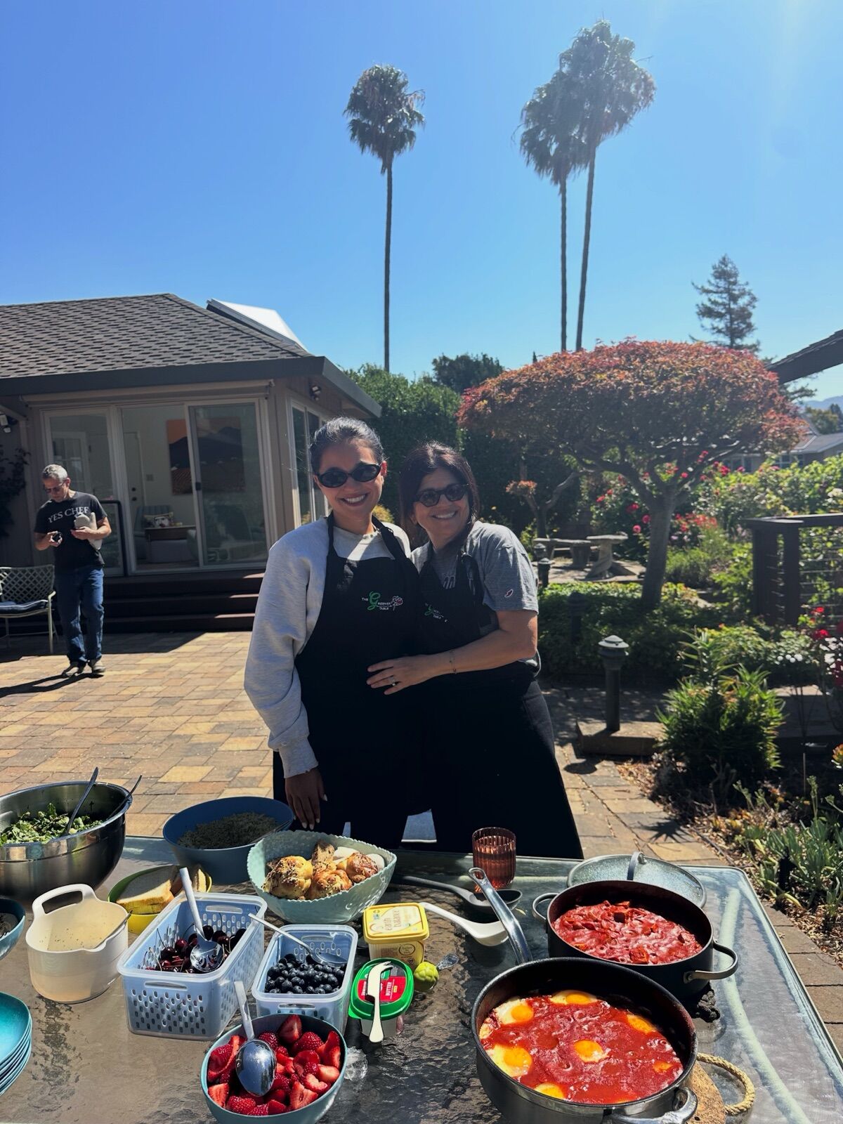 Two hosts behind pans of shakshuka at an outdoor table under the palms